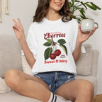White heavyweight relaxed-fit T-shirt featuring a vintage-style botanical illustration of red cherries with green leaves and retro red script reading "Fresh Picked Cherries" and "Sweet & Juicy" on a plain background — Studio DeLuz graphic tee.