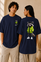 Navy oversized unisex T-shirt featuring a 3D cartoon raccoon in a lime green puffer jacket, black cap, purple sunglasses, and white sneakers — displayed front (small chest graphic) and back (large center print) on two models, Studio DeLuz.