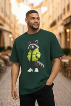 Forest green oversized unisex T-shirt with a large center-front 3D cartoon raccoon graphic wearing a lime green puffer jacket, black cap, purple sunglasses, and white sneakers — worn on a warm European cobblestone street, Studio DeLuz.
